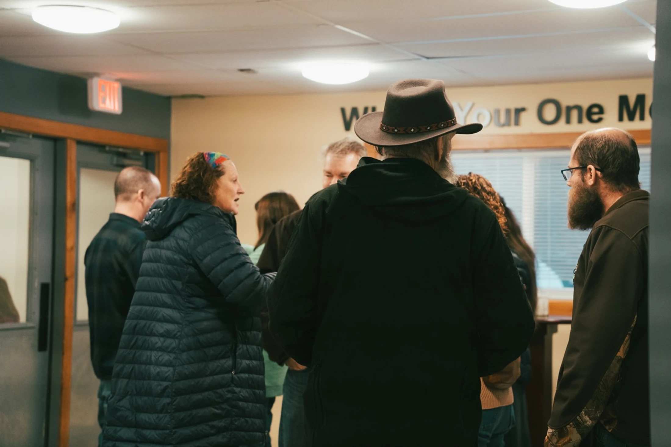 A group of casually dressed people stand talking indoors. The view is from behind a man in a black hoodie and a brown hat, who is part of the group.